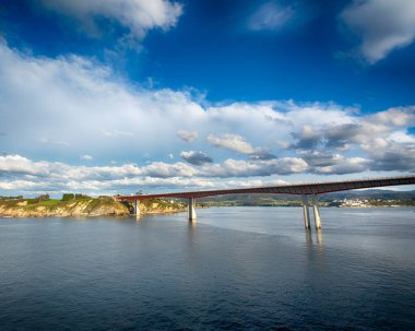 Mavi deniz ve bulutlarla dolu gökyüzü üzerinde uzun bir köprü