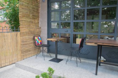 Küçük bir kafenin önündeki boş masa ve sandalye. Sandalyede renkli bir yastık. Hasır ekran ve demir doğrama sergisi.