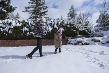 Genç bir kadın ve küçük kızı evlerinin karla kaplı bahçesinde yürüyorlar. Ayak izleriyle bir şeyler çizmeye çalışıyorlar. Kalp Şekli.