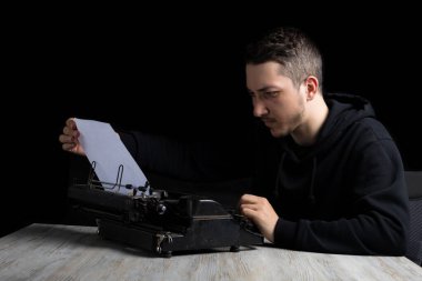 Siyah kapüşonlu genç adam daktiloda daktilo kullanıyor, siyah arka planda, gösterişsiz.
