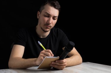 Young handsome man in black clothes with microphone, notebook and pen, on black background in low key clipart
