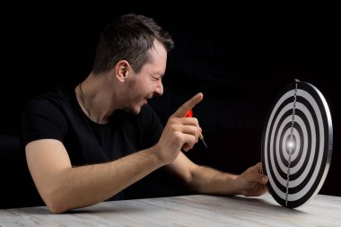 Gözleri kapalı bir adam elinde bir dart oku tutuyor. Hedefin sevinç jesti olarak başparmaklar yukarı.