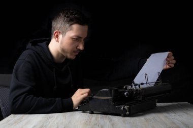 Siyah kapüşonlu genç adam daktiloda daktilo kullanıyor, siyah arka planda, gösterişsiz.