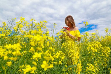 Sarı elbiseli, parlak sarı arka planda, çiçek açan kolza tohumu tarlasında genç ve güzel bir kadın. Doğayla bütünlük kavramı