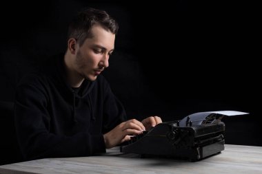 Young handsome man in black hoodie is typing on a typewriter, on a black background in low key clipart