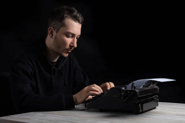 Young handsome man in black hoodie is typing on a typewriter, on a black background in low key