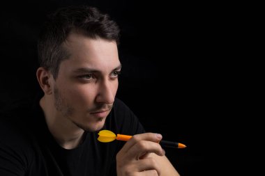 Siyah elbiseli genç yakışıklı bir adam elinde dart uçlu bir kalem tutuyor. Alçak bir anahtarın siyah arka planında. Ne pahasına olursa olsun zafer