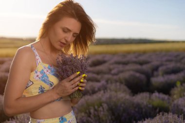 Renkli elbiseli güzel ve güzel genç bir kadın gün batımında lavanta tarlasında yürüyüp rahatlıyor.