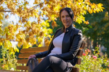 Siyah deri ceketli, 30-40 yaşlarında esmer bir kadın parkta bir bankta dinleniyor. Metin veya yazıtlar için kopyalama alanı olan arkaplan.