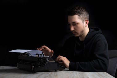 Siyah kapüşonlu genç adam daktiloda daktilo kullanıyor, siyah arka planda, gösterişsiz.
