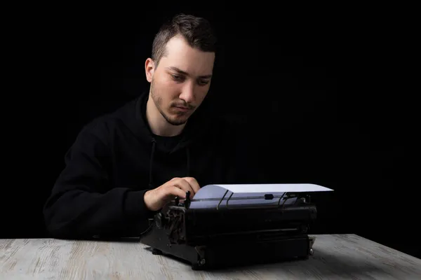 Νεαρός Όμορφος Άντρας Μαύρη Κουκούλα Δακτυλογραφεί Μια Γραφομηχανή Μαύρο Φόντο — Φωτογραφία Αρχείου
