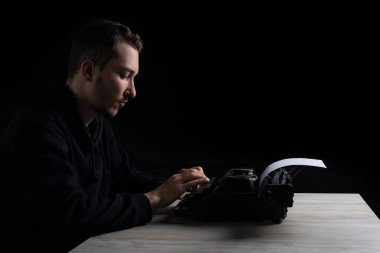 Siyah kapüşonlu genç adam daktiloda daktilo kullanıyor, siyah arka planda, gösterişsiz.