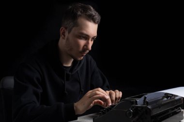 Siyah kapüşonlu genç adam daktiloda daktilo kullanıyor, siyah arka planda, gösterişsiz.