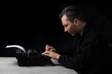 Siyah kapüşonlu genç adam daktiloda daktilo kullanıyor, siyah arka planda, gösterişsiz.