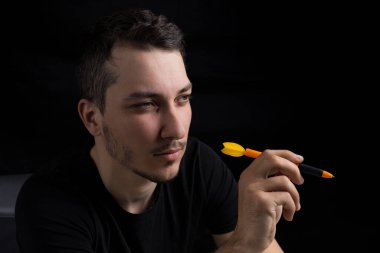 Siyah elbiseli genç yakışıklı bir adam elinde dart uçlu bir kalem tutuyor. Alçak bir anahtarın siyah arka planında. Ne pahasına olursa olsun zafer