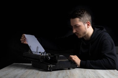 Siyah kapüşonlu genç adam daktiloda daktilo kullanıyor, siyah arka planda, gösterişsiz.