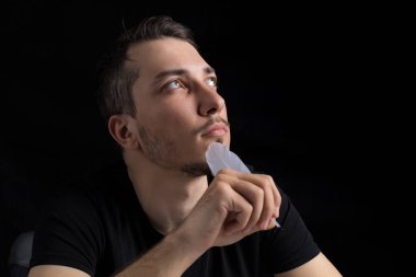 Siyah giysili, kaz tüyü giymiş yakışıklı bir genç, siyah bir arka planda dalgın ve romantik bir şekilde görünür. Metin için kopyalama alanı