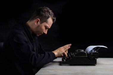 Siyah kapüşonlu genç adam daktiloda daktilo kullanıyor, siyah arka planda, gösterişsiz.