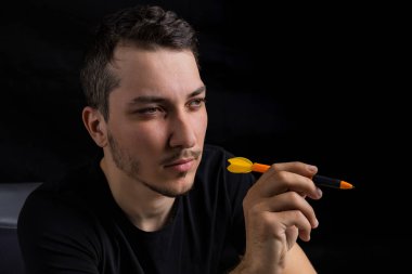 Siyah elbiseli genç yakışıklı bir adam elinde dart uçlu bir kalem tutuyor. Alçak bir anahtarın siyah arka planında. Ne pahasına olursa olsun zafer