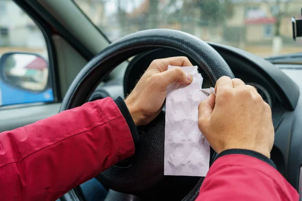 The driver opens a package of pills against a blurred background of the steering wheel in the car. The use of pharmacological drugs for medical purposes while driving. Selective focus, toned