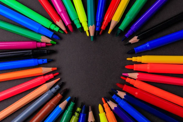 Pencils and felt-tip pens form a copy space in the shape of an abstract heart on a black background symbolizing the blackboard.