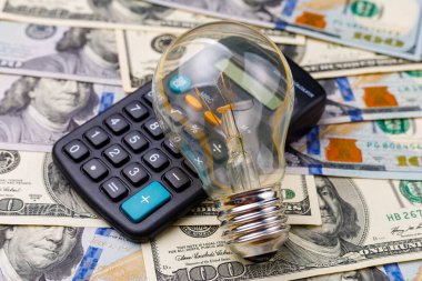 Hesap makinesi ve doların bulanık arka planında klasik şeffaf parlak ampul, seçici odaklanma. Enerji krizi ve elektrik enflasyonu kavramı. Artan elektrik fiyatları