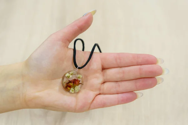Transparent pendant with flowers in the palm of a female hand. Homemade epoxy resin jewelry. Rare interesting unusual hobby. Background with selective focus and copy space for text