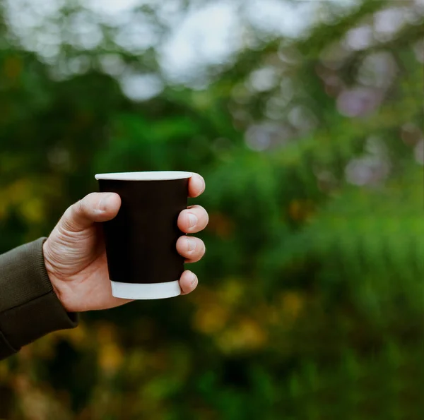 남자는 나무가 우거진 배경에서 빈 종 이 컵을 들고 손을 잡고 있다. 가을의 생활 양식. 커피 블랙 컵. 복사 공간 — 스톡 사진