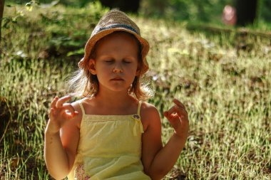 The child meditates outdoors. Moments of childhood. Copy space. Grass background. Girl 5 years old in a hat and yellow dress.