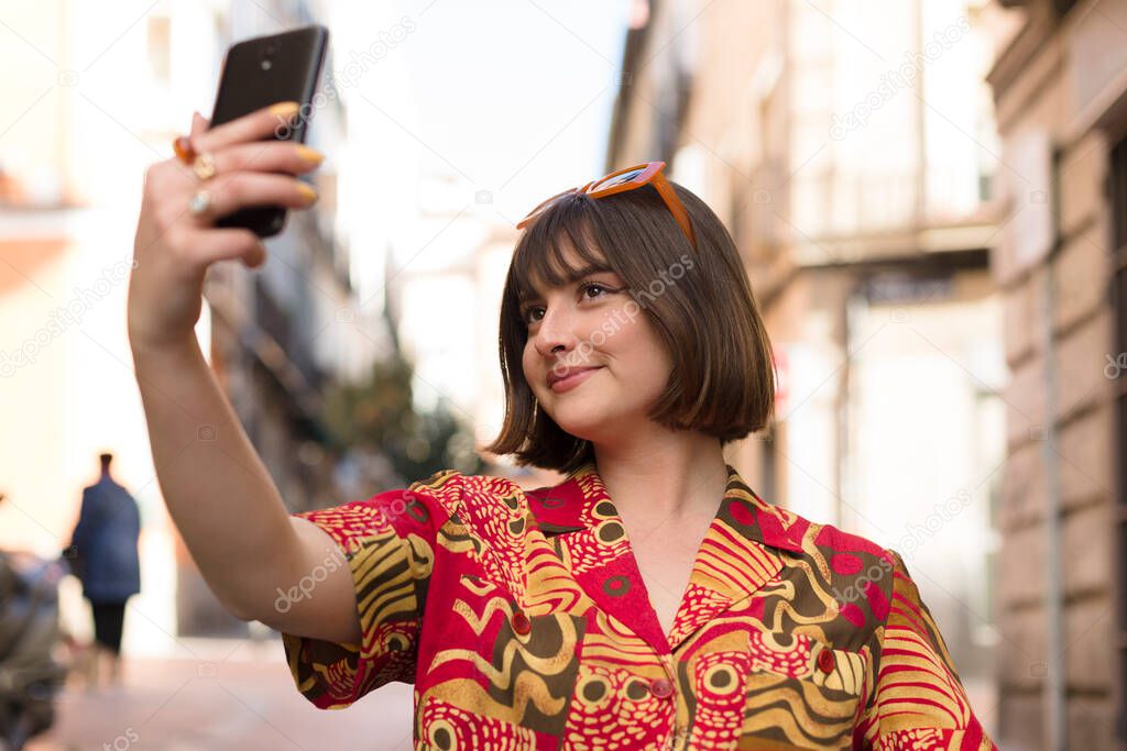Smiling young caucasian woman with short hair taking a photo of herself with a mobile phone outdoors.