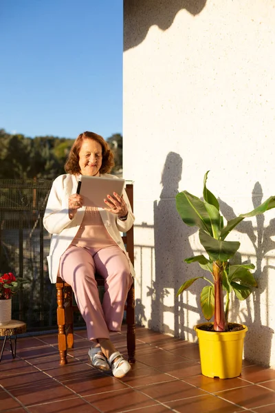 Elderly Caucasian woman using a portable digital tablet on the terrace of her house. Concept of technology, communications and elderly people. Space for text.