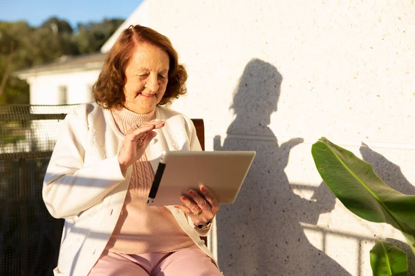 Elderly Caucasian woman making a video call from a digital tablet on the terrace of her house. Concept of elderly people and new technologies. Space for text.