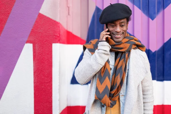 Young adult African American man talking on phone outdoors. He is standing next to a colorful wall. Space for text.