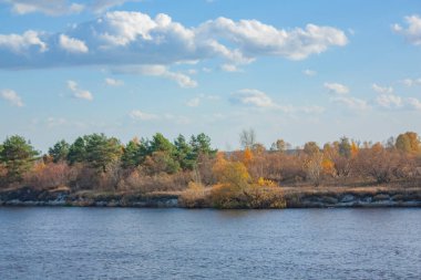 Mavi gökyüzünde güzel bulutlar. Parlak güneşli bir günde sonbahar manzarası. Nehrin karşı kıyısındaki sarı ve yeşil ağaçlar ve çalılar.