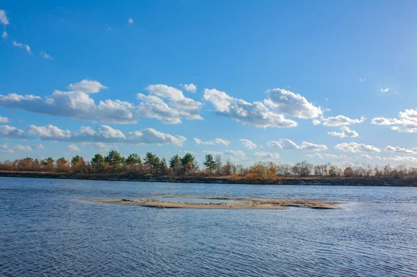 Mavi gökyüzünde güzel bulutlar. Parlak güneşli bir günde sonbahar manzarası. Nehrin karşı kıyısındaki sarı orman.
