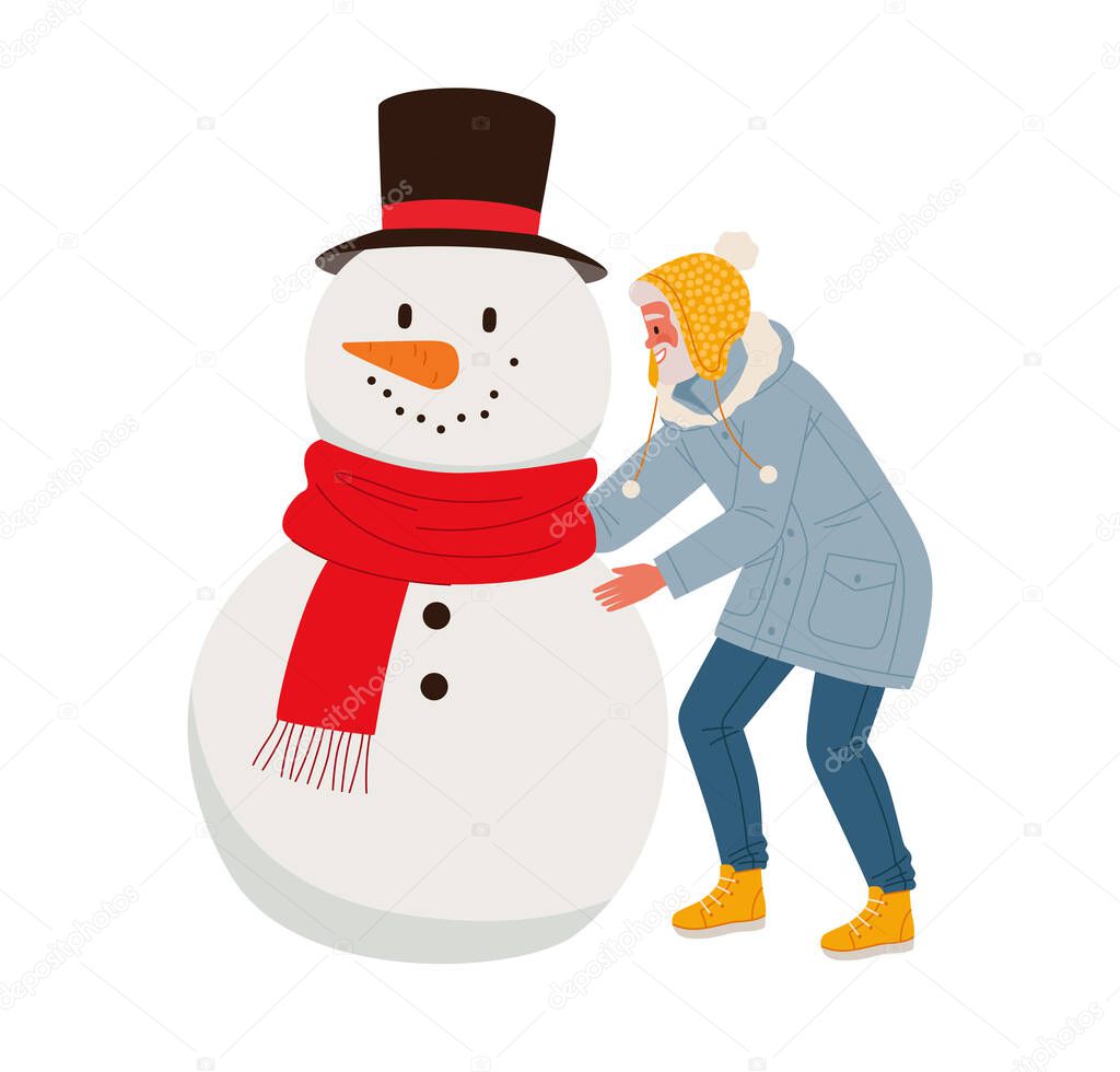 Stylishly dressed man or old man with a beard in a long jacket with a hood makes a snowman wearing a red scarf. Fun outdoor entertainment for the elderly