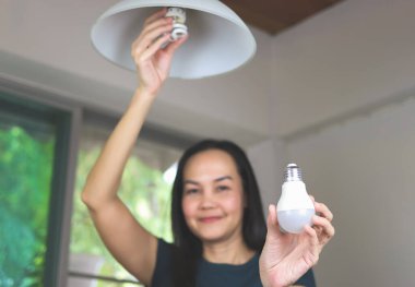 Neşeli Asyalı kadın evinde ampul değiştiriyor. LED ampulü floresan ile değiştir, LED ampulü kameraya göster. güç tasarruf kavramı.