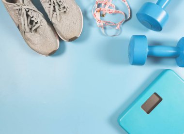 Top view or flat lay of blue weight scale, blue dumbbells, gray sneakers and measuring tape on blue background with copy space. Sports and weight loss concept.