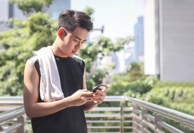 Portrait of Asian young man wearing sportswear, standing outdoor on walkway with city building background, seriously reading message on mobile phone.
