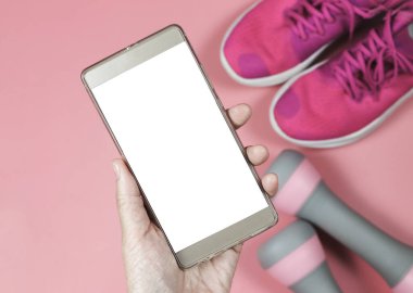 Close up image of mobile phone in hand, blank white screen with pink sneakers, pink dumbbells, pink background. Mobile app for active lifestyle.