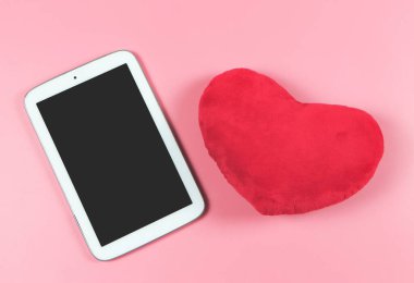 Bilgisayar tabletinin üst görüntüsü ve pembe arka planda kırmızı kalp şeklindeki yastığın fotokopi alanı. Sevgililer Günü ve teknoloji konsepti.