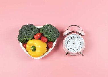 Üst ya da düz sebze kapsicum, brokoli ve domates pembe arka planda kalp şeklinde tabak, klasik çalar saat, sağlıklı yaşam tarzı ve aralıklı oruç kavramı.