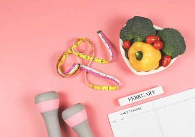 Alışkanlık takip kitabının üst görüntüsü, tahta takvim şubat, gri pembe halterler, ölçüm bandı ve kalp şeklindeki sebzeler, pembe arka planda fotokopi alanı. Sağlıklı yemek, yaşam tarzı konsepti.