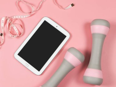 Üst ya da düz siyah ekranlı tablet, pembe halterler ve pembe arkaplandaki mezura. Spor ve kilo verme konsepti.