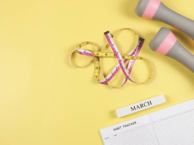 Üst ya da düz alışkanlık takip kitabı, ahşap takvim marşı, gri pembe dambıllar ve sarı arka planda fotokopi alanı ile ölçüm bandı. Kişisel gelişim ve aktif yaşam tarzı kavramı.