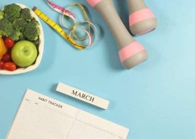 Alışkanlık takip kitabının üst görüntüsü, ahşap takvim marşı, gri pembe halterler, bant ve sebzeler mavi arka planda mavi arka planda kalp şeklinde plaka. Sağlıklı yemek, yaşam tarzı konsepti.