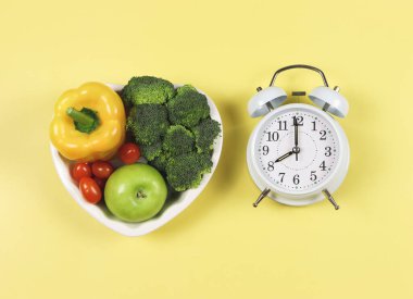 Sebze, kapsika, brokoli, domates ve elmanın sarı zemin üzerindeki kalp şeklinde tabakta beyaz antika çalar saat, sağlıklı yaşam tarzı ve aralıklı oruç kavramı..