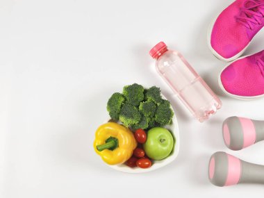 Capsicum, brokoli, domates ve elma kalp şeklinde tabakta, pembe gri halterler, pembe spor ayakkabılar veya spor ayakkabıları ve beyaz zemin üzerinde bir şişe su. sağlıklı yaşam tarzı.