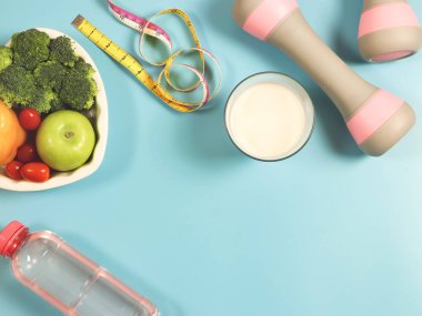 Kalp şeklinde tabakta sebze ve meyve manzarası, bir şişe su, mezura, pembe halterler ve mavi arka planda süt. Sağlıklı yaşam tarzı kavramı.