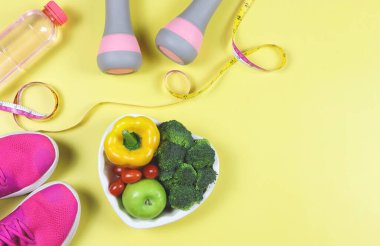 Üst ya da düz sebze yatağı ve kalp şeklinde tabakta meyve, bir şişe su, mezura, pembe halterler ve sarı arka planda pembe spor ayakkabılar..